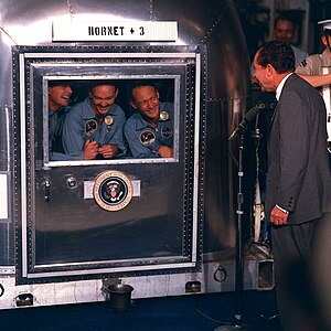 Photo of the three crew members smiling at the President through the glass window of their quarantine chamber. President Nixon is standing at a microphone, also smiling.